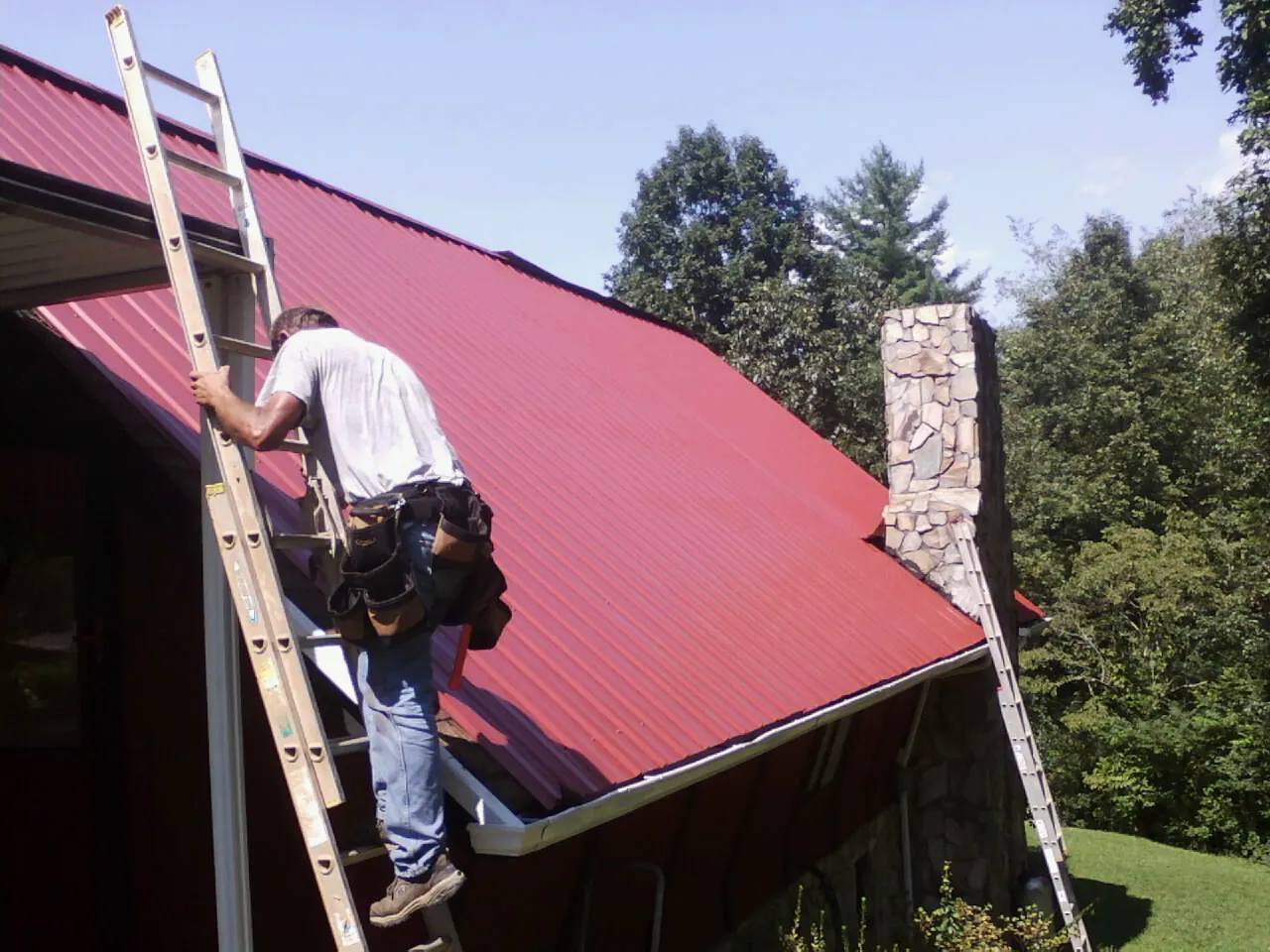 Roofer on a ladder beside a red metal roof and stone chimney — Soffit & Fascia Repair by Roofing Brothers in Pikeville